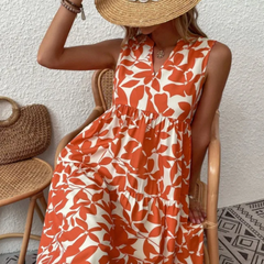 A woman sits on a chair in a sleeveless, orange and white leaf-patterned dress made from lightweight breathable fabric. She holds a wide-brimmed straw hat over her face and accessorizes with bracelets, a necklace, and a woven bag.