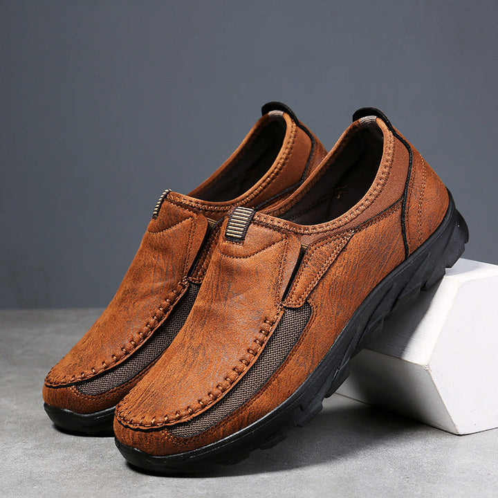 A pair of brown Men Leather Casual Shoes with black rubber soles are displayed, one shoe propped up on a white block. These breathable loafers feature visible stitching and elastic side panels for a comfortable fit and easy wearing.