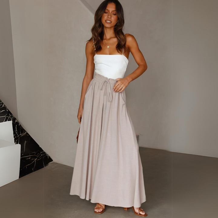 A woman stands indoors wearing a white strapless top and a long, flowy wide-leg beige skirt with a tie at the waist. She has wavy brown hair and sandals. The background is neutral with minimal decor.