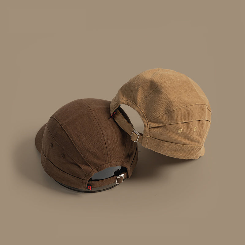 Two Japanese-style peaked caps in brown and tan are placed side by side on a plain beige background, both displaying the adjustable strap with metal buckles at the back.
