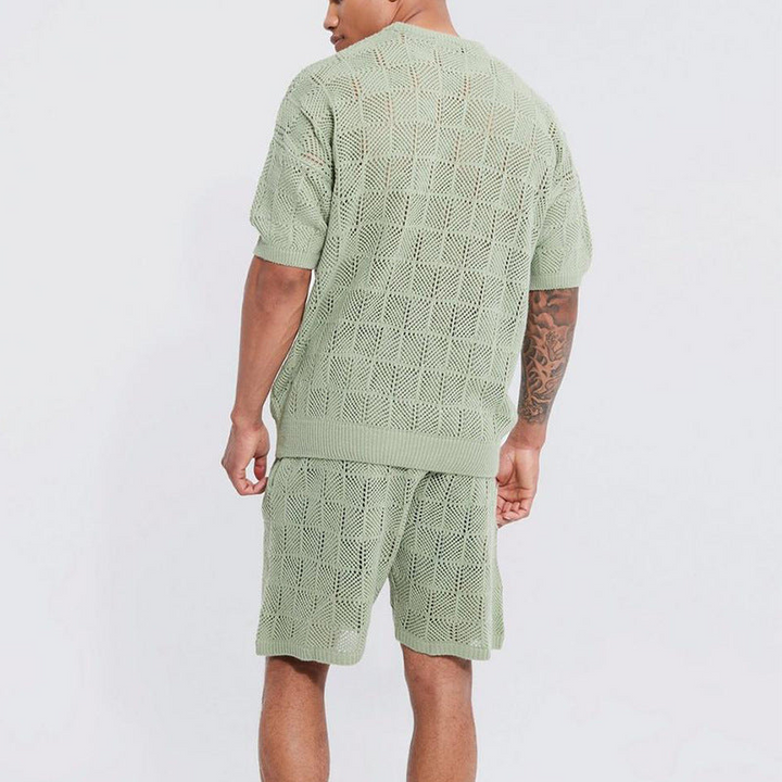 A man stands with his back to the camera, wearing a light green knit men's 2 piece set featuring a casual short sleeve shirt and matching shorts. The outfit showcases a checkered, semi-see-through pattern—perfect for summer style.