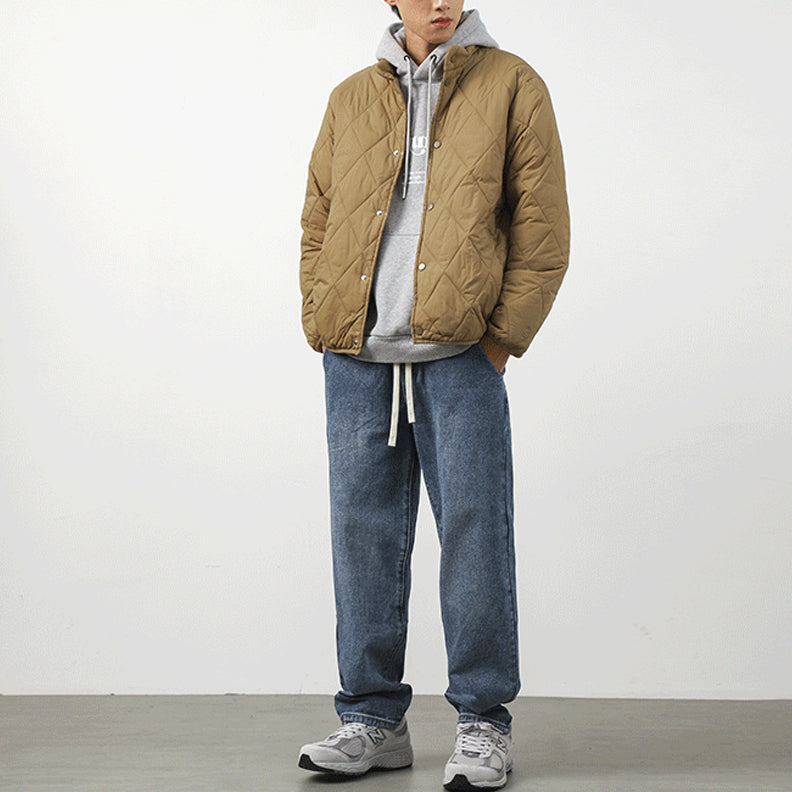 A person stands against a plain white background wearing a tan diamond-stitched pattern padded jacket over a gray hoodie, blue jeans, and gray sneakers. Their hands are in their jacket pockets, and their face is partially obscured by the hood.