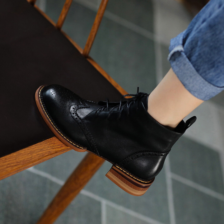 An urban trendsetter sits on a wooden chair in rolled-up jeans, showing off genuine leather brogue boots with decorative perforations and a stylish brown sole.