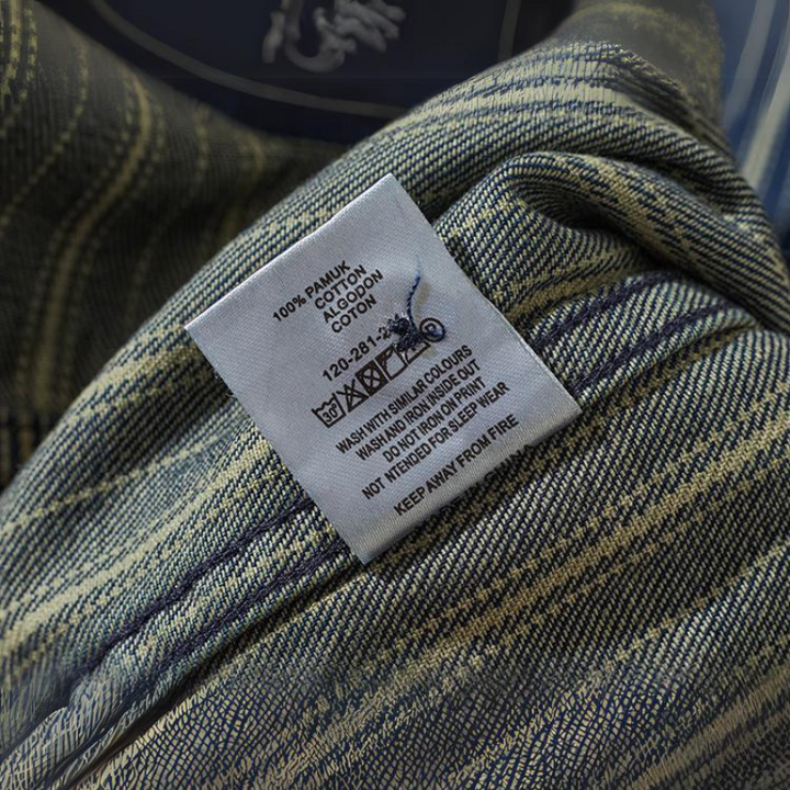 A close-up of a clothing care label on a relaxed-fit button-down plaid fabric, showing washing and ironing instructions and noting the material is 100% cotton. The tag also advises to keep the garment away from fire.