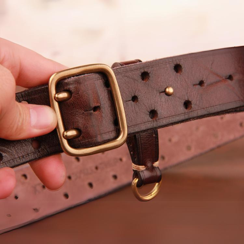 A hand holds a vintage leather belt with brass hardware, including a rectangular buckle and a small ring. The premium leather belt features multiple adjustment holes and visible stitching.