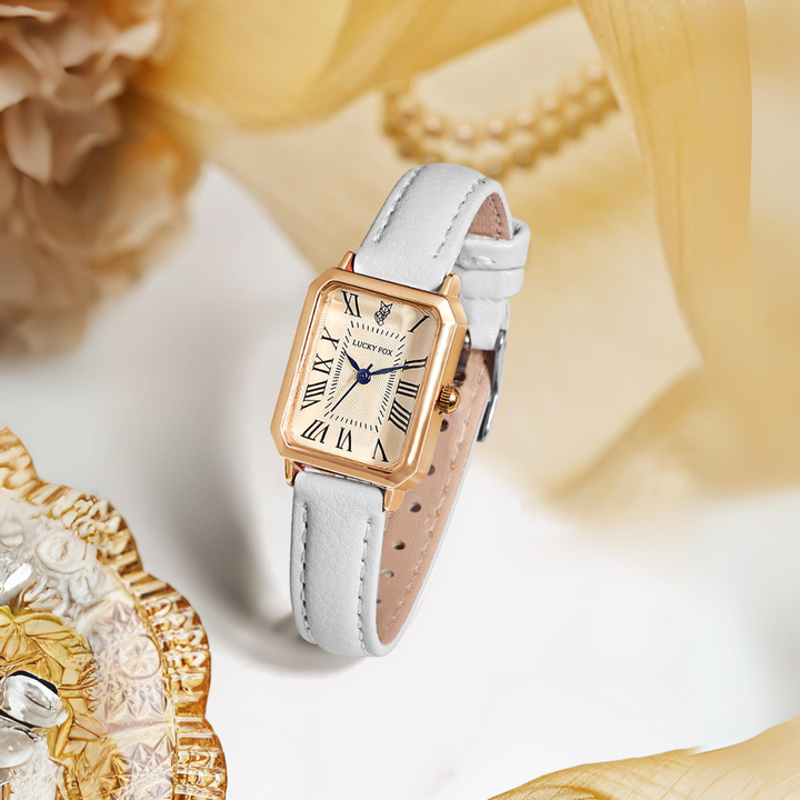A rectangular face watch with a gold case, white genuine leather strap, and Roman numerals rests on a light surface, surrounded by soft golden fabric, pearls, and decorative glassware.