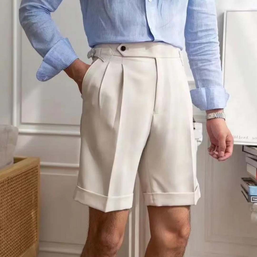 A person wearing a light blue dress shirt and high-waisted pleated shorts stands indoors with one hand in their pocket, channeling an old money aesthetic through refined details and a tailored fit.