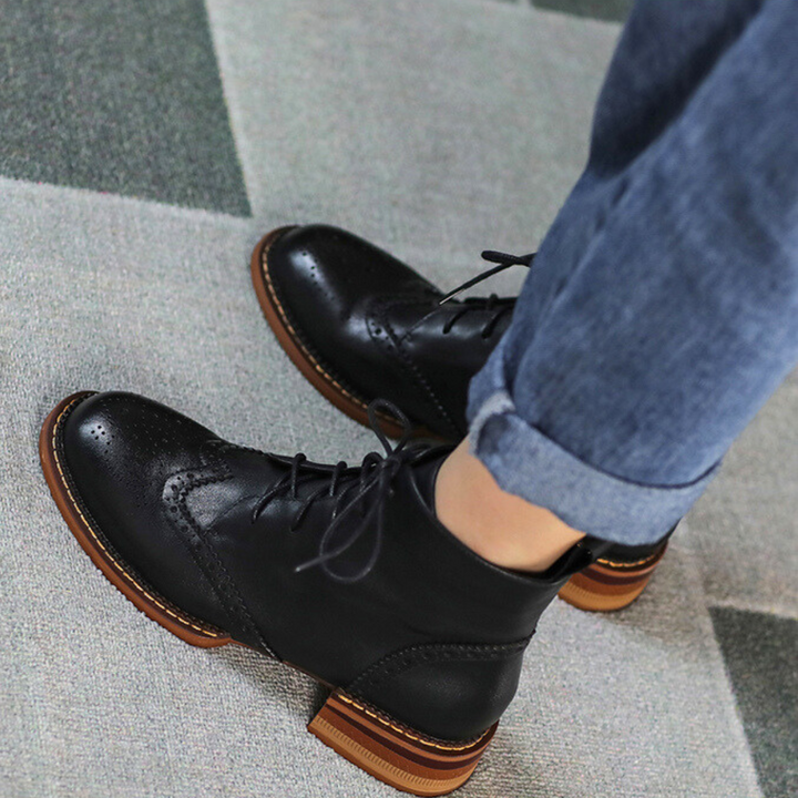 An urban trendsetter stands on a patterned carpet, wearing dark blue rolled-up jeans paired with classic black leather brogue boots featuring brown soles.