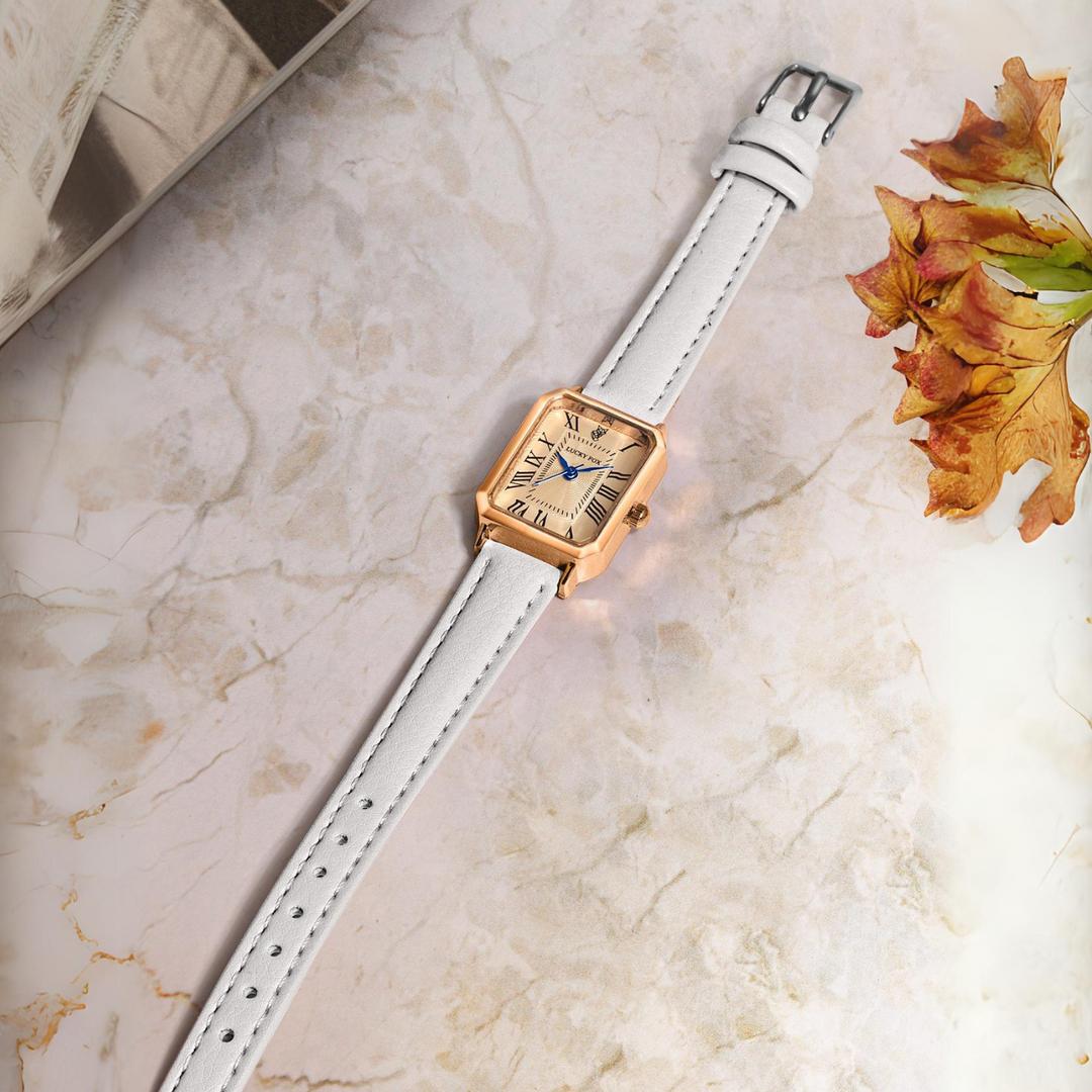 A gold rectangular face watch with a genuine leather strap rests on a marble surface. The Roman numeral watch features blue hands, while autumn leaves and a book are partially visible nearby.