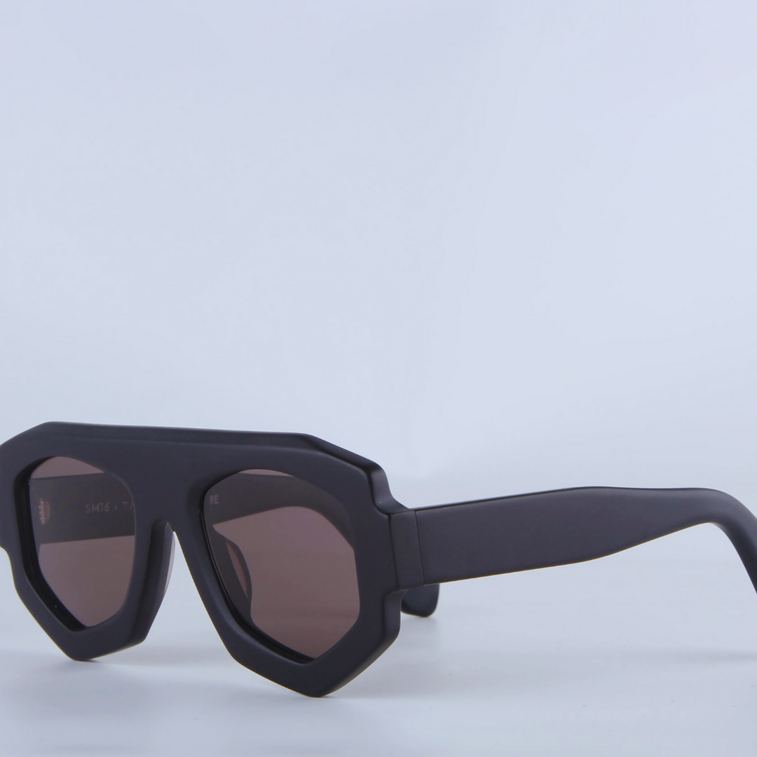 Black geometric sunglasses with thick acetate frames and dark, UV400 protection lenses are displayed against a plain, light background. The sunglasses are positioned at an angle, showcasing their bold geometric frame design.