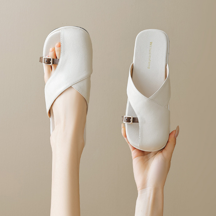 A pair of chic slides with white crisscross straps and small side buckles, held up by two hands against a neutral beige background—the perfect addition to your summer wardrobe.