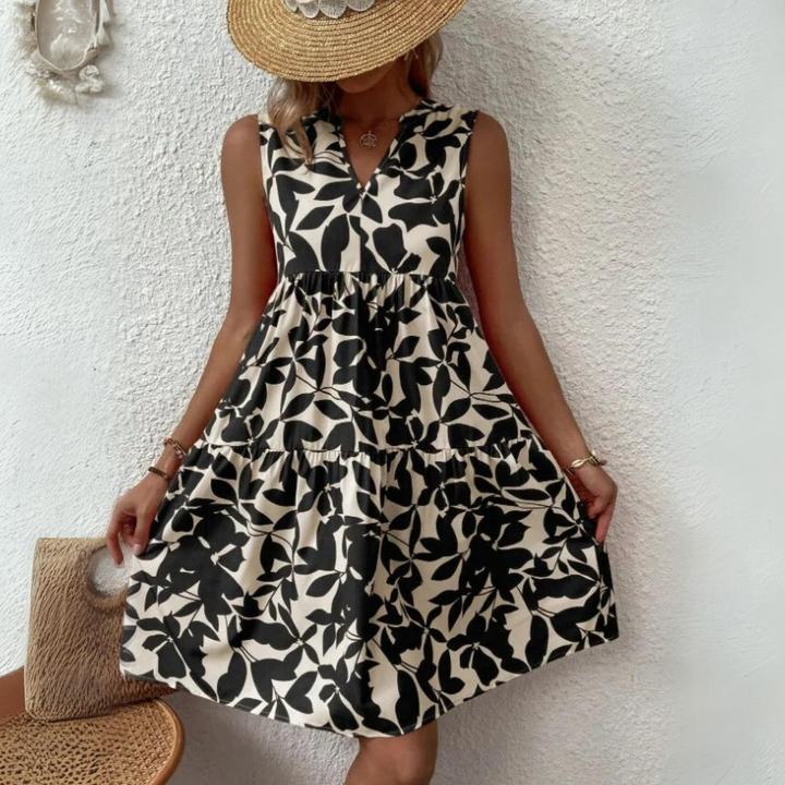 A woman in a lightweight, breathable sleeveless dress with a black and white leaf pattern holds the sides of her skirt. She wears a wide-brimmed straw hat, partially covering her face, and stands against a textured white wall.