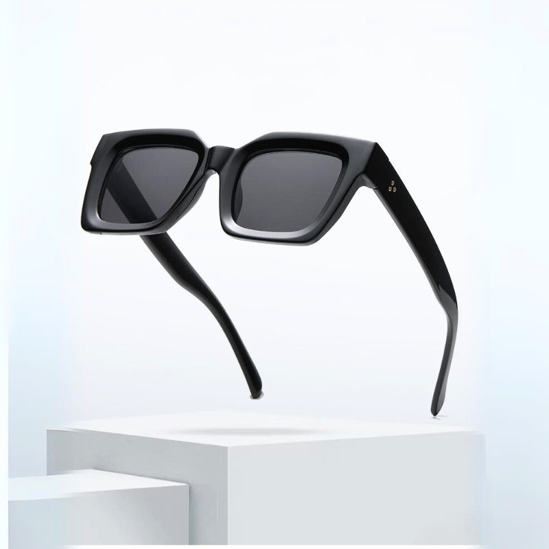 A pair of modern, black square-framed sunglasses with thick acetate frames and dark lenses, offering UV protection, float above a minimalist white pedestal against a light background.