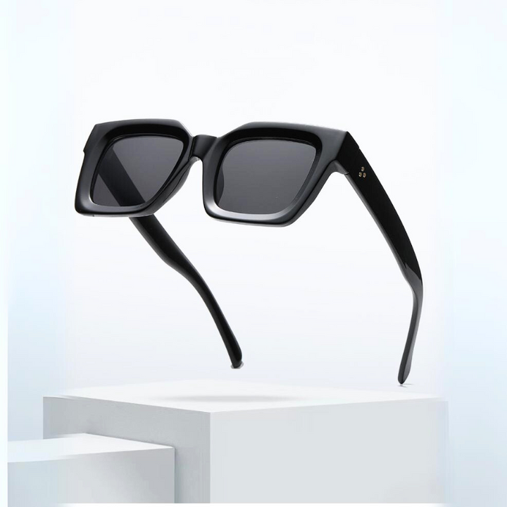 A pair of modern, black square-framed sunglasses with thick acetate frames and dark lenses, offering UV protection, float above a minimalist white pedestal against a light background.