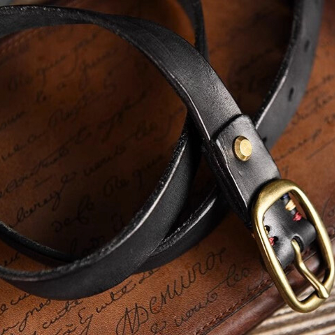 A black leather belt with a brass buckle is laid on a brown surface featuring cursive handwritten text. The coiled belt, with sturdy stitching and a metal fastening rivet, evokes timeless country fashion perfect for Southern ladies.
