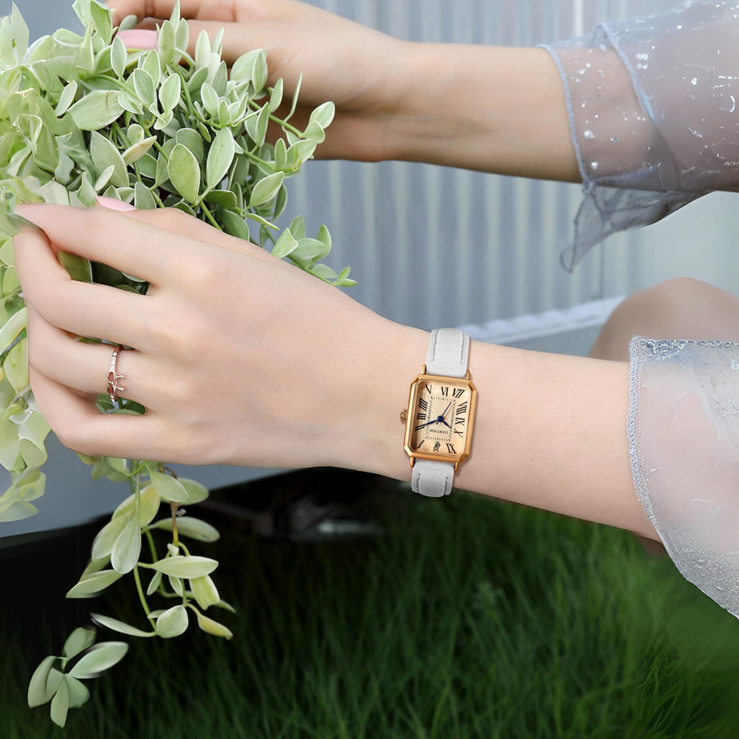 A person wearing a gold rectangular face watch with a white genuine leather strap gently holds a green leafy plant. The person is dressed in a sheer, light blue, long-sleeved top and has a delicate ring on their finger.