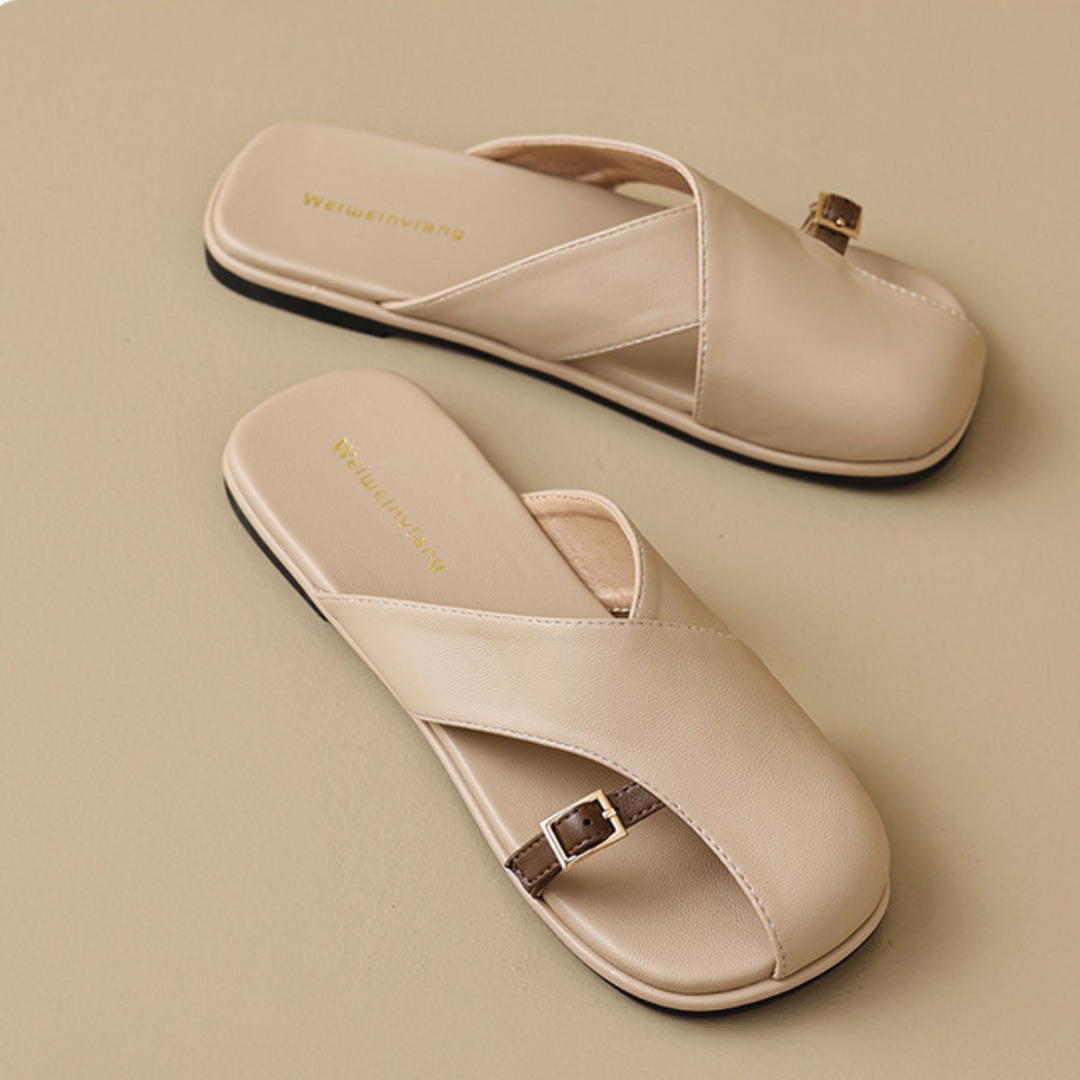 A pair of chic beige leather slides with crisscross straps, a small buckle on the side, and flat black soles, displayed on a neutral background—perfect for elevating your summer wardrobe.
