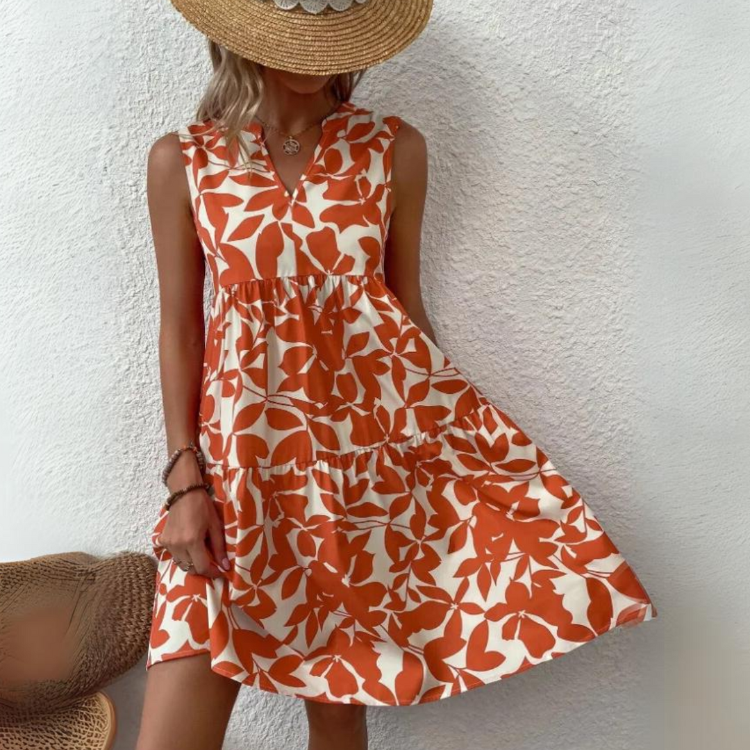 A woman in a tan sunhat and a sleeveless, orange and white floral dress—crafted from lightweight breathable fabric—stands against a white textured wall, holding out the hem of her summer wardrobe essential. Her face is mostly hidden by the hat.