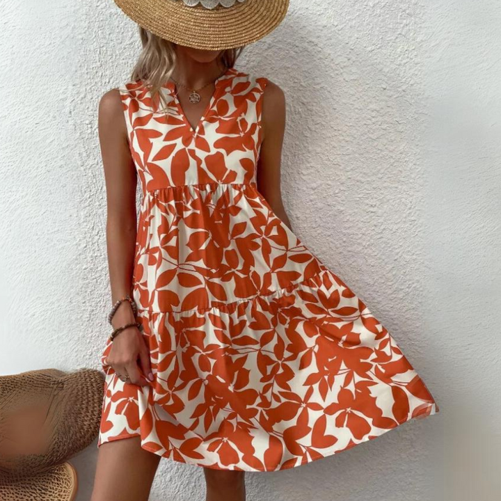 A woman in a tan sunhat and a sleeveless, orange and white floral dress—crafted from lightweight breathable fabric—stands against a white textured wall, holding out the hem of her summer wardrobe essential. Her face is mostly hidden by the hat.