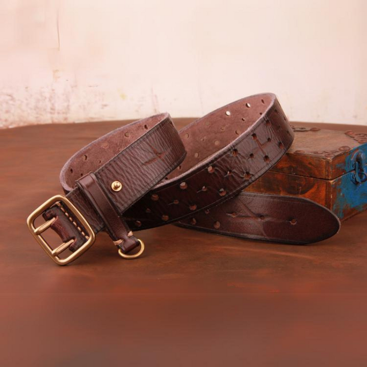 A vintage leather belt with a brass buckle is coiled on a wooden surface next to a rustic box. The premium leather belt features a textured finish and visible perforations along its length.