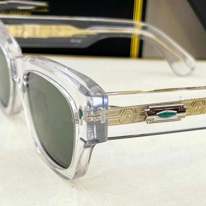 Clear-framed retro sunglasses with dark lenses, featuring a decorative gold and green geometric design on the arms, rest on a light surface with a black and yellow box in the background. Enjoy stylish UV400 protection all day.