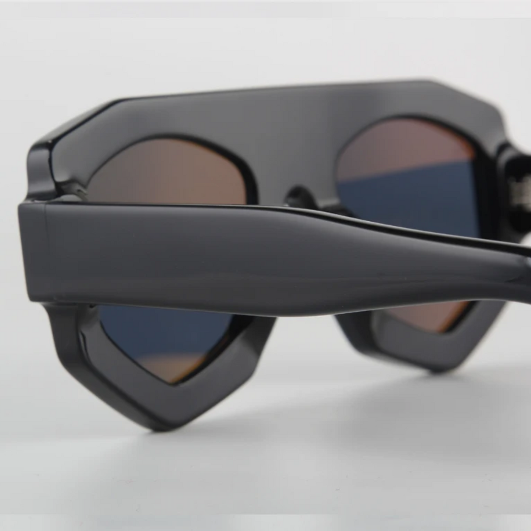 Close-up view of angular, geometric black sunglasses with dark lenses and thick acetate side arms, set against a plain white background. The image highlights the sharp-edged geometric frame and bold design.