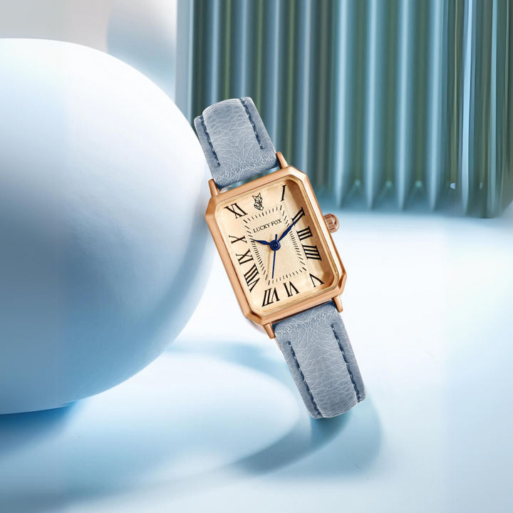 A rectangular face watch with a gold case, light blue genuine leather strap, Roman numeral hour markers, and blue hands is displayed against a soft blue and white minimalist background.