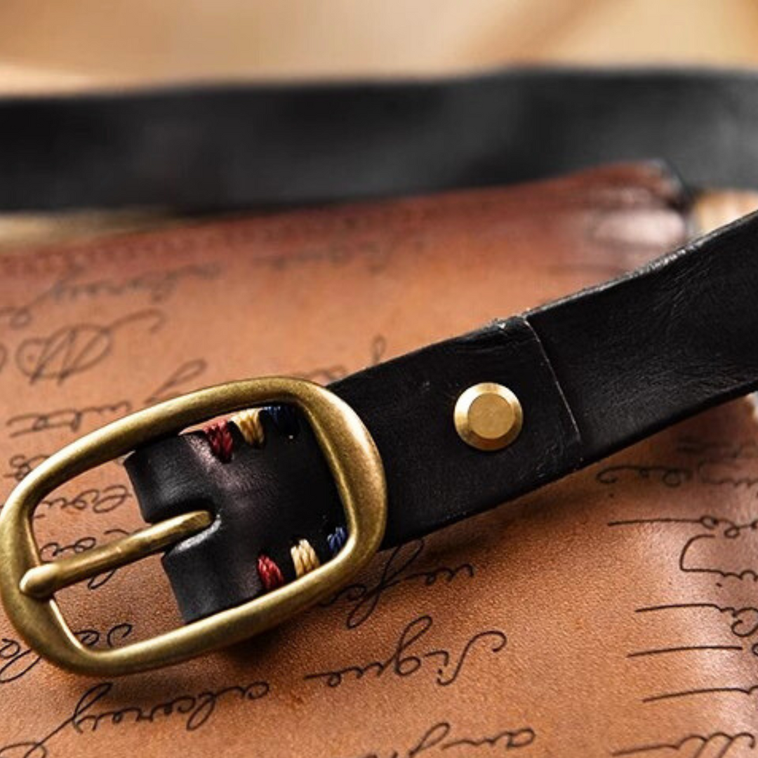 A close-up of a black leather belt with a brass buckle rests on a brown surface decorated with elegant cursive writing. The strap features stitching in multiple colors, adding a touch of country fashion next to the buckle.