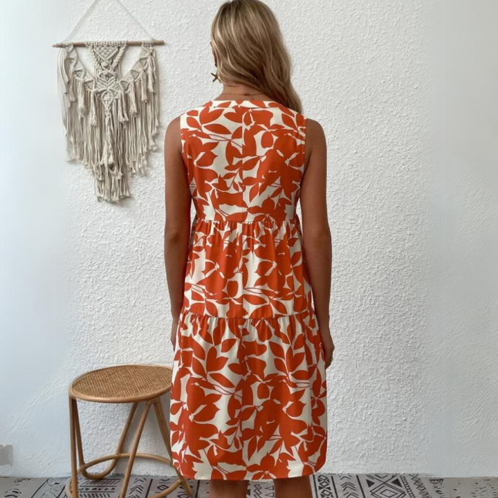 A woman with long blonde hair stands facing a white textured wall, wearing a sleeveless orange and white leaf-patterned dress—an ideal addition to any summer wardrobe. A woven stool and a macramé wall hanging are visible beside her.