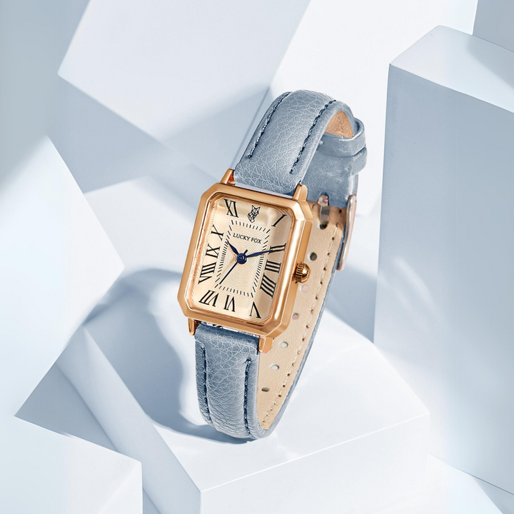 A gold rectangular face watch with a cream dial, black Roman numerals, and a light blue genuine leather strap is displayed on white geometric blocks with soft lighting.