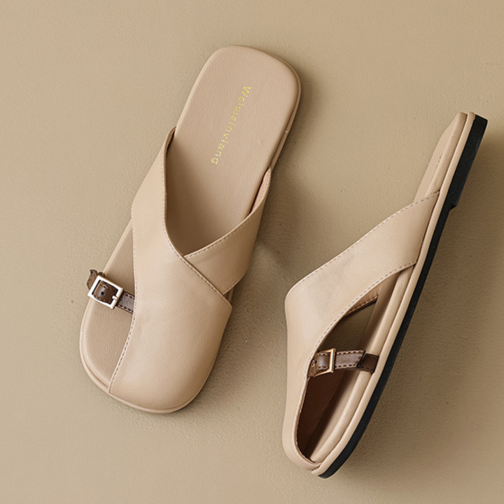 A pair of chic leather slides in beige with crisscross straps, brown buckle accents, and black soles—perfect for elevating your summer wardrobe, displayed on a neutral beige background.