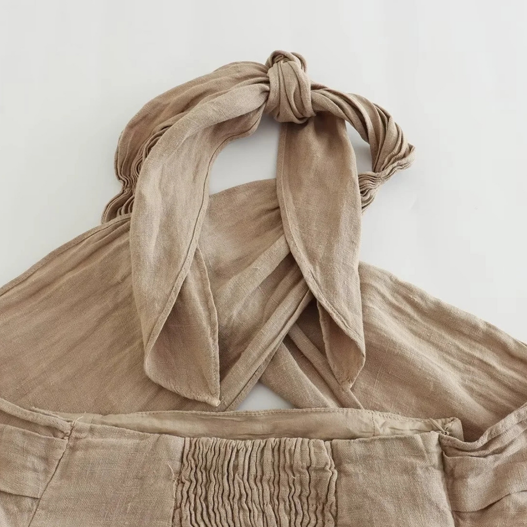 A close-up of a beige, linen halter top with a chic halter collar and wide straps tied in a knot at the neckline, displayed on a white surface. The fabric has a slightly wrinkled texture and visible gathered details.