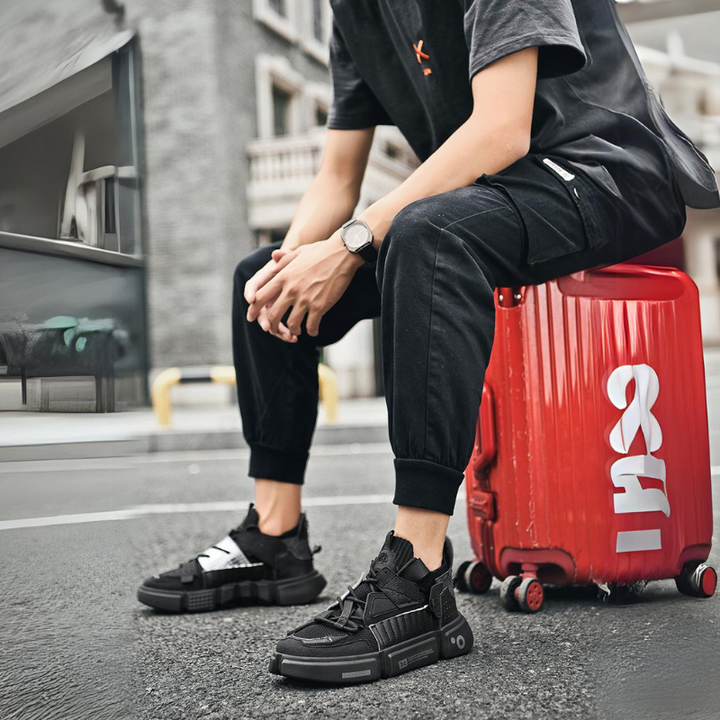 A person in black joggers and lightweight breathable sneakers sits on a red suitcase on a wet street, with urban buildings in the background. The persons face is not visible.