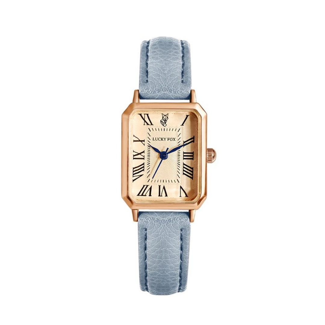 A rectangular face watch with a gold case, cream dial, blue Roman numerals, blue hands, and a small fox logo at the top. It features a textured light blue genuine leather strap for added elegance.