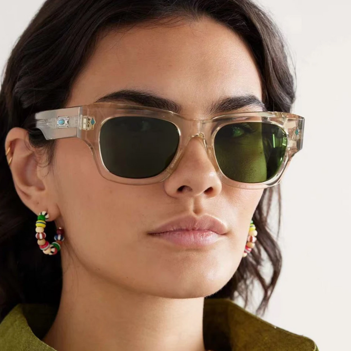 A woman with dark wavy hair wears vintage-inspired sunglasses with green lenses and colorful beaded hoop earrings, looking straight ahead with a neutral expression.