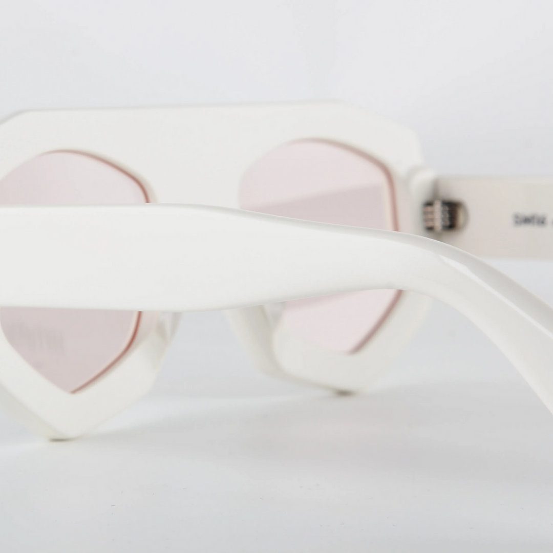 Close-up of white acetate sunglasses with pink-tinted lenses and a geometric frame, focusing on the temple and frame against a plain white background.