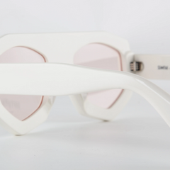 Close-up of white acetate sunglasses with pink-tinted lenses and a geometric frame, focusing on the temple and frame against a plain white background.