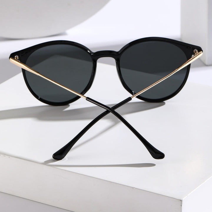 Black round sunglasses with dark lenses, gold temples, and black earpieces are displayed on a white surface, featuring UV protection and anti-glare properties. The arms are crossed at the back, casting a shadow.