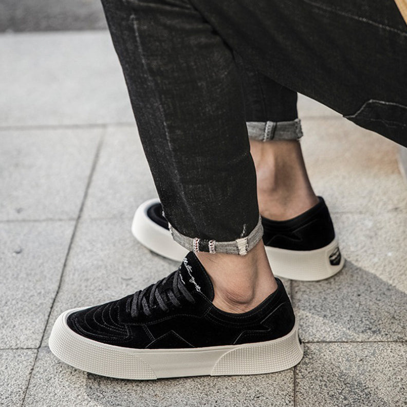 A person wearing black lace-up sneakers with comfortable cushioning and white soles, paired with rolled-up black jeans, sits on a tiled outdoor surface.
