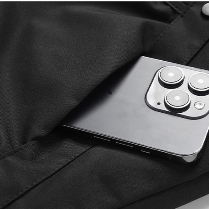 A silver smartphone with three camera lenses peeks out from the pocket of black pants, next to a windproof jacket with adjustable cuffs.