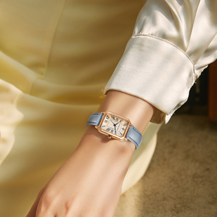 A person wearing a cream-colored satin blouse sits with their arm resting, showcasing a rectangular face watch with a blue genuine leather strap and an elegant Roman numeral dial.