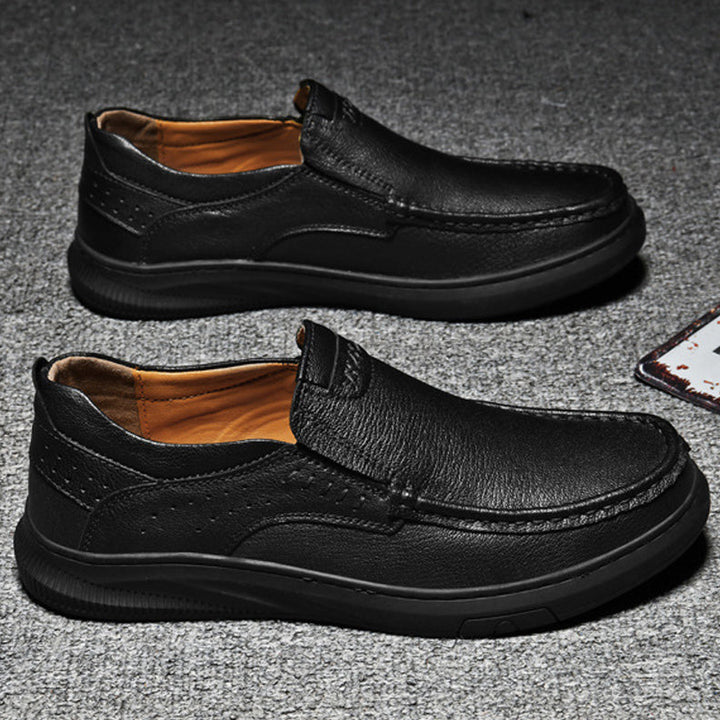A pair of Men's Cowhide Leather Slip-On Shoes with soft, textured soles and brown inner lining, displayed side by side on a gray textured surface.