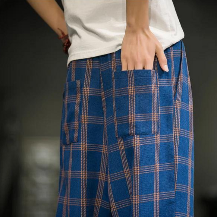 A person wearing loose-fitting blue plaid cotton joggers with large front pockets rests their hand on one pocket. Only the lower torso and part of one arm are visible, and the person wears a white shirt, channeling Tokyo streetwear vibes.