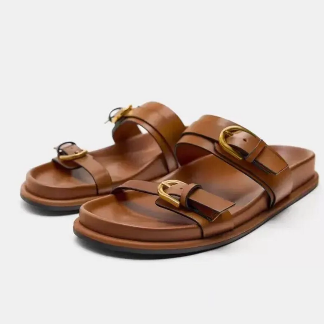 A pair of brown leather sandals with a slip-on style and two wide straps with gold buckles, featuring a flat sole, black outsoles, and leather upper, displayed on a white background.