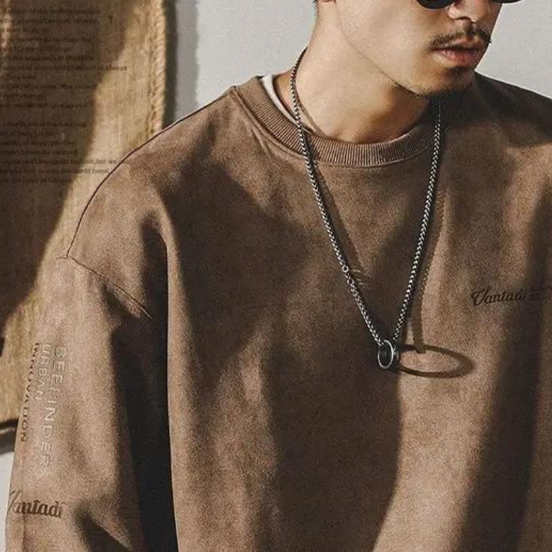 A man wearing sunglasses, a silver chain necklace, and a relaxed fit crewneck sweatshirt stands against a neutral background. Only his upper body is visible and he is looking slightly downward.