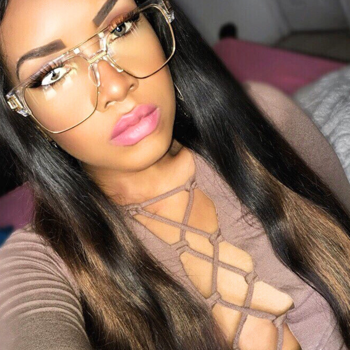 A woman with long straight hair wears large geometric sunglasses with UV400 protection and a brown top with a crisscross lace-up front. She has bold eyebrows and pink lipstick, posing indoors on a bed with pink covers.