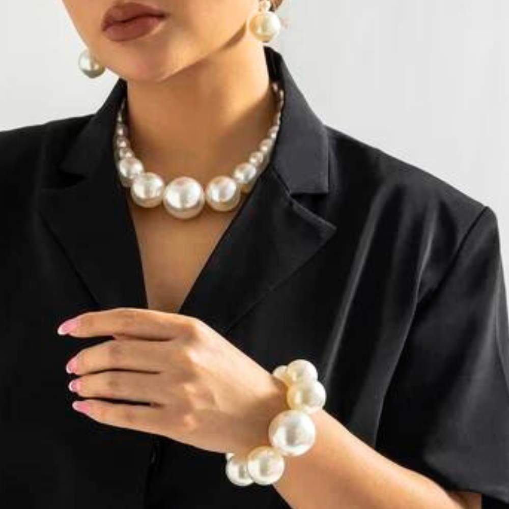A woman wearing a black blazer is shown from the shoulders down, accessorized with a pearl jewelry set featuring large pearl earrings, a matching necklace, and a chunky bracelet—elegant statement jewelry paired with light pink nails.