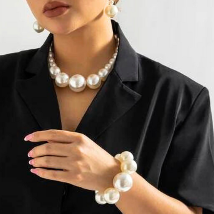 A woman wearing a black blazer is shown from the shoulders down, accessorized with a pearl jewelry set featuring large pearl earrings, a matching necklace, and a chunky bracelet—elegant statement jewelry paired with light pink nails.