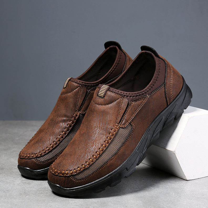 A pair of brown Men Leather Casual Shoes with black rubber soles, decorative stitching, and mesh side panels for a comfortable fit, displayed against a gray background with one shoe propped on a white block.