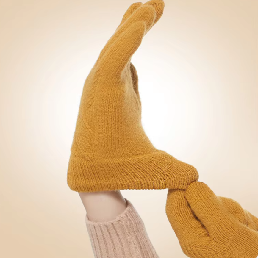 Two hands wearing luxurious, mustard yellow knitted gloves touch at the fingertips, forming an arch against a light beige background. One cashmere blend glove is lightly grasped at the wrist by the other hand.
